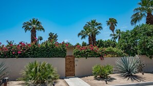 A gated entrance with a wooden door, surrounded by palm trees and flowering bushes. - ATOMIC-MOD HOUSE in prime Palm Desert/next to El Paseo! (Palm Desert)