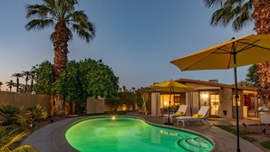 A swimming pool with a curved edge, surrounded by palm trees and a patio area with lounge chairs and an umbrella. - ATOMIC-MOD HOUSE in prime Palm Desert/next to El Paseo! (Palm Desert)
