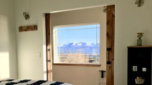 A bedroom with a large window offering a mountain view, a bed with a striped bedspread, a wooden wardrobe, and a small table with a vase.