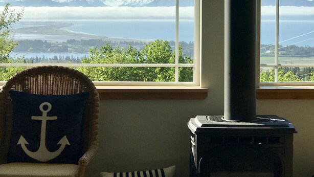A room with a large window offering a view of mountains and a lake. There is a wicker chair with a cushion and a wood-burning stove.