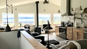 A spacious living area with a high ceiling, large windows offering a view of the outdoors, a kitchen with modern appliances, and a cozy seating area with a fireplace.
