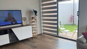 Un soggiorno moderno con televisione, pavimento in legno, un armadietto bianco, una scala e una porta vetrata scorrevole che dà su un patio con mobili da esterno.