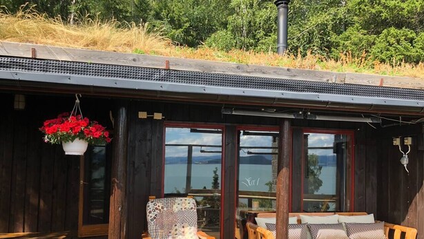 Une cabane en bois dotée d’une grande fenêtre donnant sur un plan d’eau, d’une terrasse en bois, d’une jardinière suspendue et d’un coin salon avec des coussins.