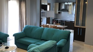 Een moderne woonkamer met een blauwgroene bank, een glazen salontafel en een eethoek, met een keuken op de achtergrond.