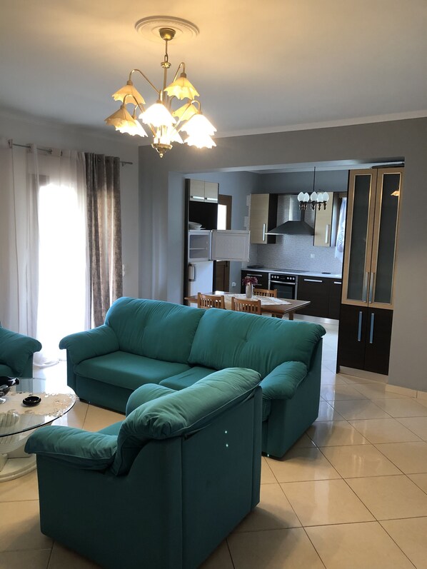 A modern living room with a teal sofa, a glass coffee table, and a kitchen area in the background.