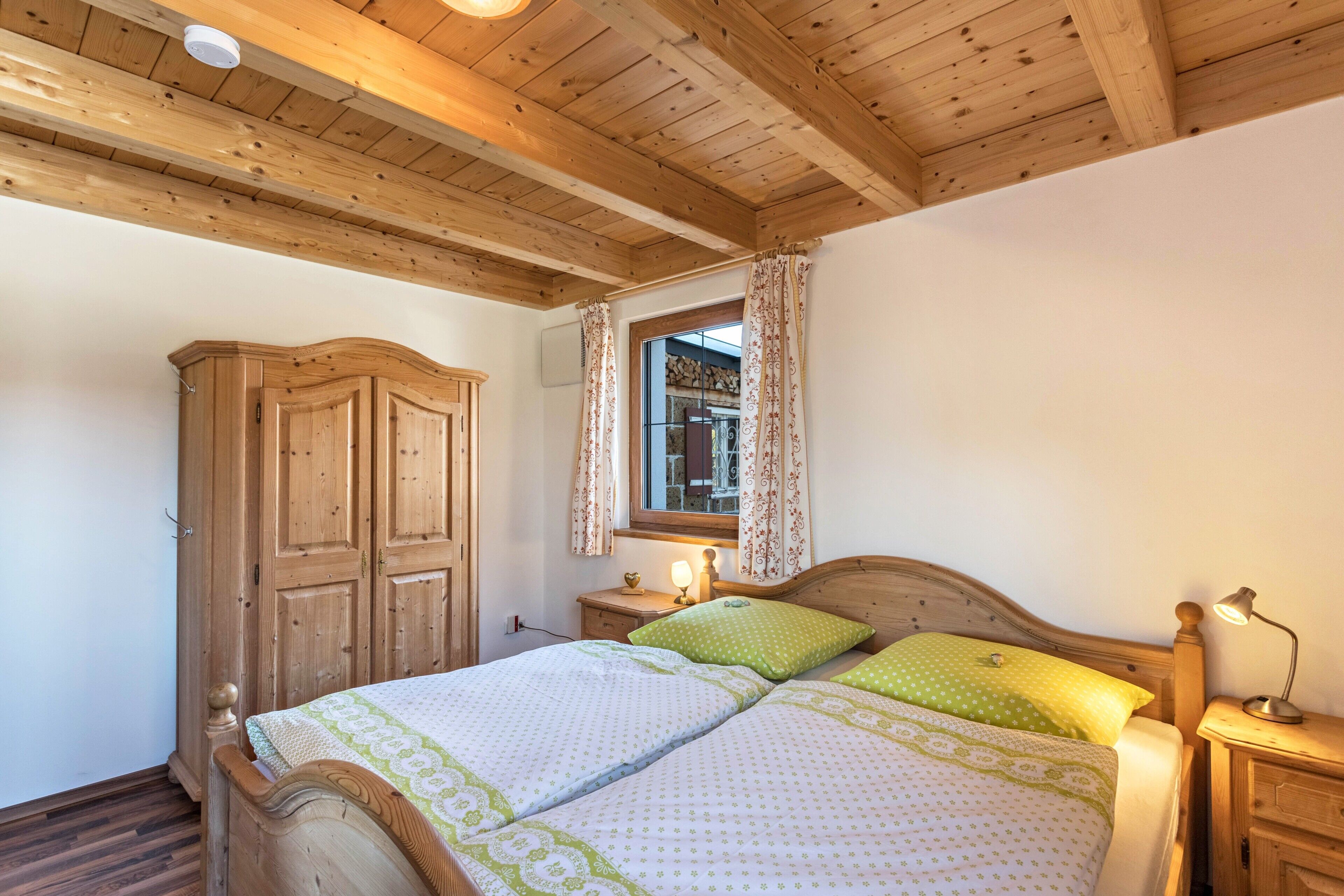 A bedroom with a wooden ceiling, a wardrobe, a bed with green and white bedding, a nightstand with a lamp, and a window with curtains.