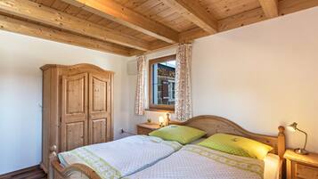 A bedroom with a wooden ceiling, a wardrobe, a bed with green and white bedding, a nightstand with a lamp, and a window with curtains.