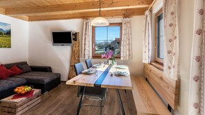 A dining area with a wooden table, chairs, a bench, a TV mounted on the wall, and a window with curtains.