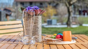A wooden table with a glass vase containing purple flowers, a glass pitcher, a cheese board with various cheeses, and a wooden bench in the background.
