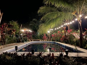 Ein Poolbereich bei Nacht mit Lichterketten, Palmen und tropischen Pflanzen.