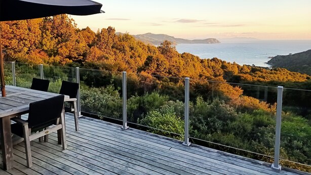 A wooden deck with a table and chairs, overlooking a coastal landscape with trees and the sea. - Little Bay Eco House - Kaka Cottage overlooking the ocean and up in the trees (Waikawau)