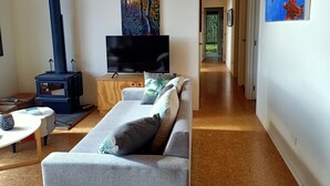 A living room with a sofa, a TV, a wood-burning stove, and paintings on the walls. - Little Bay Eco House - Kaka Cottage overlooking the ocean and up in the trees (Waikawau)