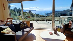A modern living room with a large glass door leading to a deck with a scenic ocean view. - Little Bay Eco House - Kaka Cottage overlooking the ocean and up in the trees (Waikawau)