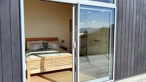 A bedroom with a bed, a nightstand, and a glass door leading to a balcony. - Little Bay Eco House - Kaka Cottage overlooking the ocean and up in the trees (Waikawau)