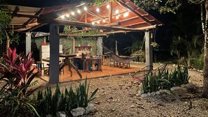 Un espacio al aire libre techado con estructura de madera, mesas, sillas y una hamaca.