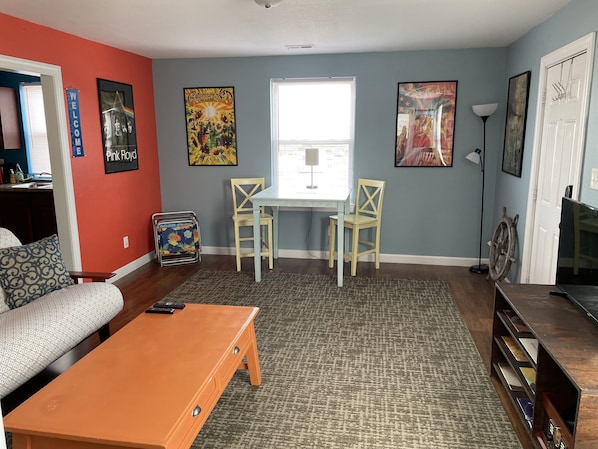 A living room with a sofa, coffee table, and a dining area with chairs. There are wall art pieces and a lamp.