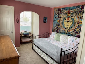 A bedroom with a bed, a wooden desk, a window with blinds, and a colorful tapestry on the wall.