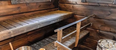 A wooden sauna with benches, a metal bowl, a wooden spoon, and a stone container.