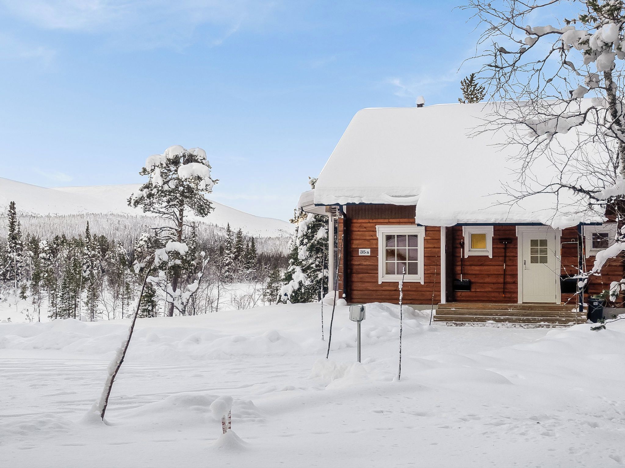 En stuga av trä med ett snötäckt tak, omgiven av ett snöigt landskap och en skog.