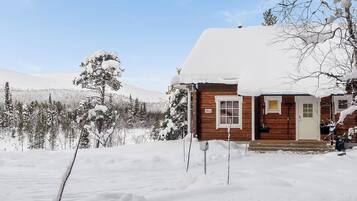 En stuga av trä med ett snötäckt tak, omgiven av ett snöigt landskap och en skog.