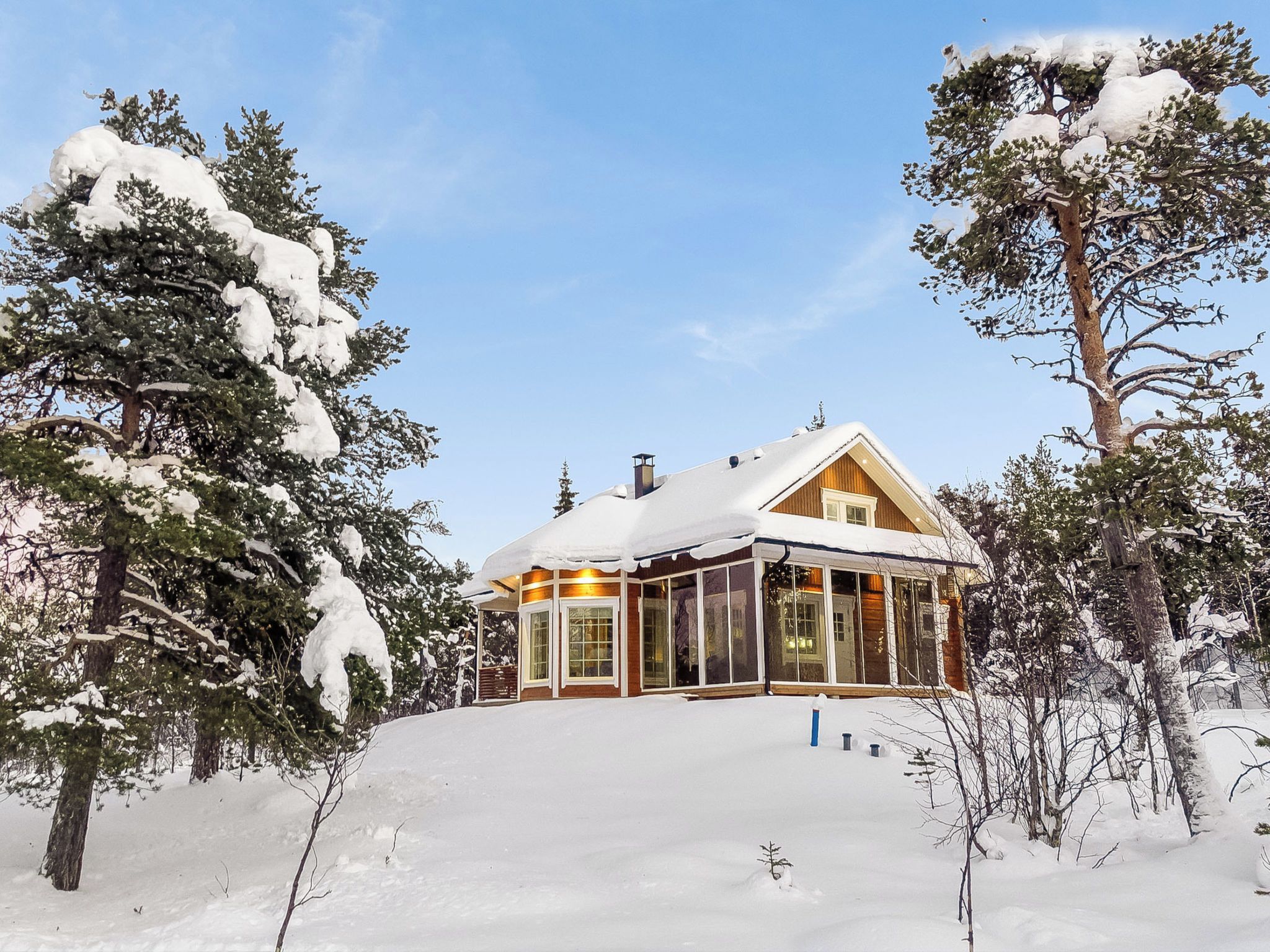 Lumipeitteinen mökki, jossa on puinen ulkoverhous, suuret ikkunat ja savupiippu.