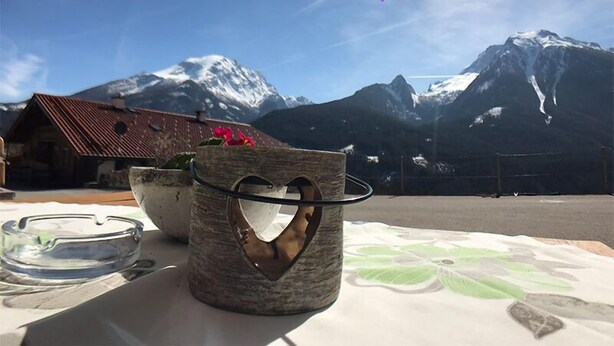 A rustic wooden table with a clear glass ashtray and a stone container with a heart-shaped cutout. In the background, a mountain with snow-capped peaks and a clear blue sky is visible.