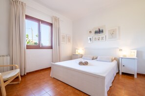 Ein Schlafzimmer mit Bett, Nachttisch, Sessel, Fenster mit Vorhängen und an der Wand befestigten Bildern.