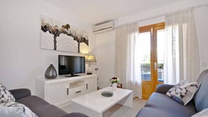 Un salon avec un téléviseur, une table basse blanche, un canapé gris et une fenêtre avec des rideaux.
