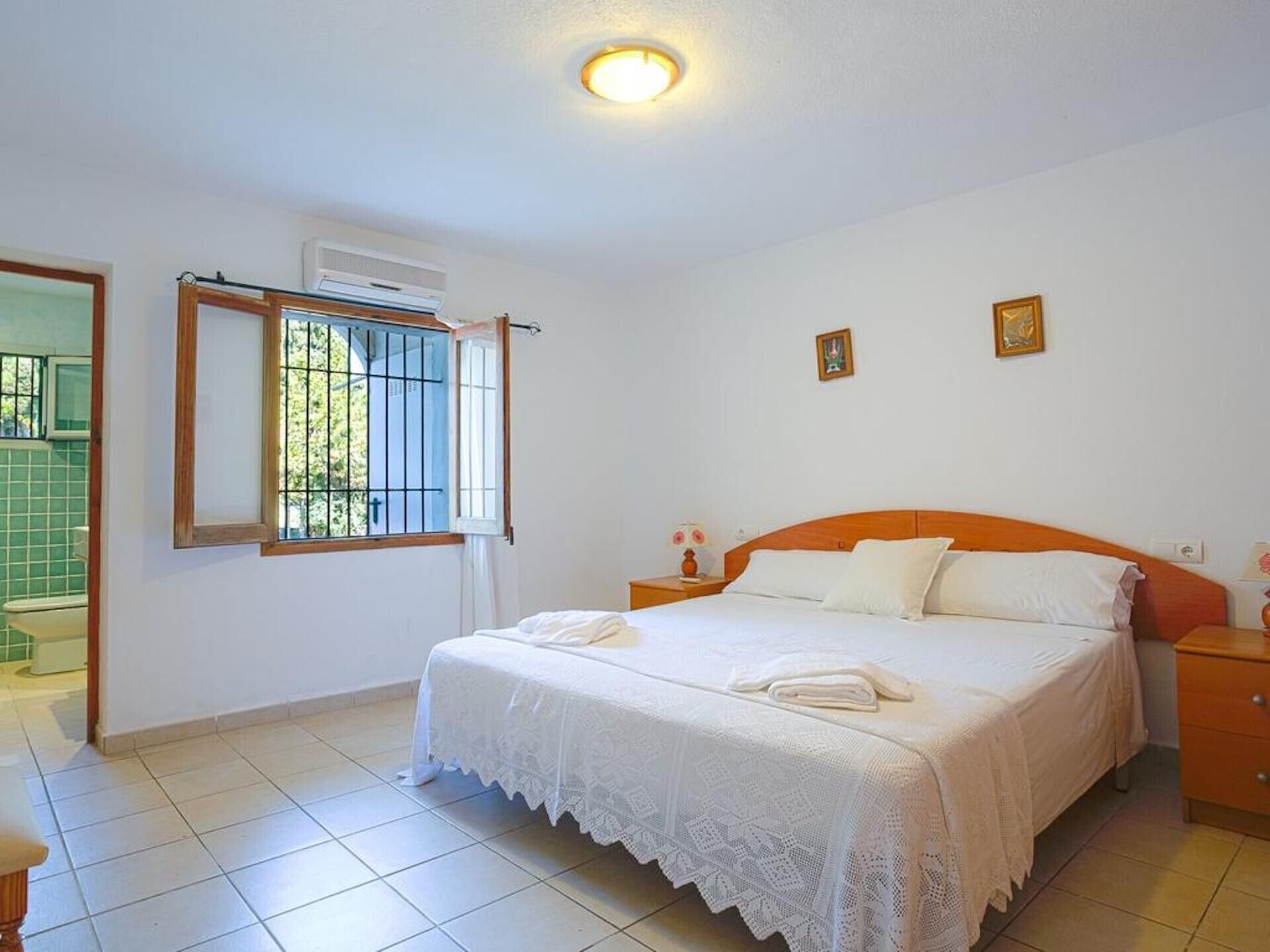 Une chambre à coucher comprenant un lit, une table de chevet, une fenêtre avec des barreaux et une porte donnant sur une salle de bain.