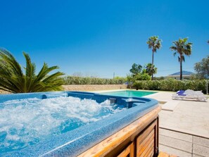 A hot tub with a clear blue pool in the background, surrounded by palm trees and a stone wall.