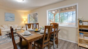 Un coin repas avec une table en bois dressée pour quatre personnes, entourée de chaises. On aperçoit un canapé et une lampe au fond, ainsi qu’une fenêtre donnant sur des arbres.