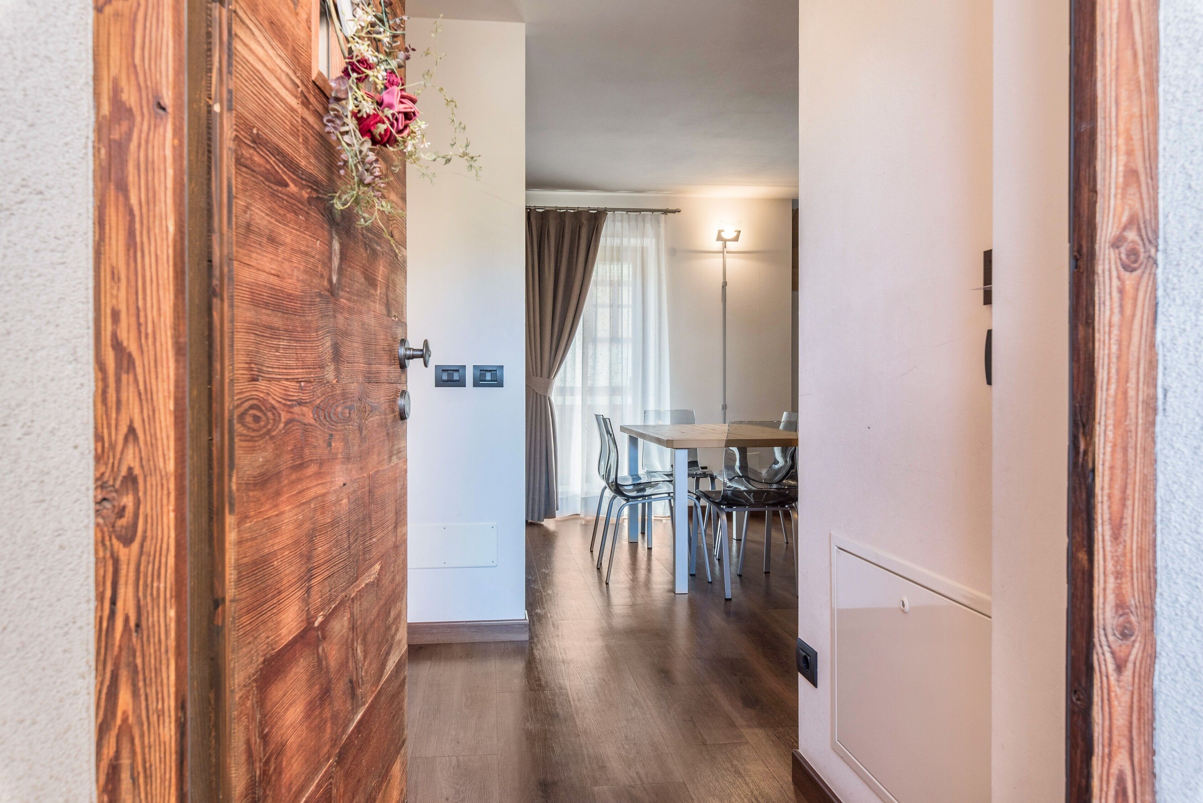 Un pasillo con una puerta de madera, un comedor con mesa y sillas, y una lámpara en la pared.