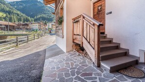 A wooden staircase leading to a door with a number plate, a stone-paved area, and a view of buildings and mountains in the background.