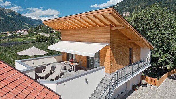 A modern two-story house with a wooden facade, a balcony with outdoor seating, and a view of a mountainous landscape. - Vacation Apartment "Sauvignon" with Mountain View, Wi-Fi, Balcony, Terrace & Garden (Meran)