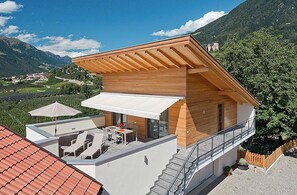 A modern two-story house with a wooden facade, a balcony with outdoor seating, and a view of a mountainous landscape. - Vacation Apartment "Sauvignon" with Mountain View, Wi-Fi, Balcony, Terrace & Garden (Meran)