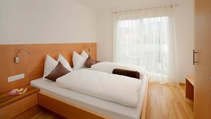 A modern hotel room with a wooden headboard, a large bed with white bedding, a bedside table, and a window with sheer curtains. - Vacation Apartment "Sauvignon" with Mountain View, Wi-Fi, Balcony, Terrace & Garden (Meran)