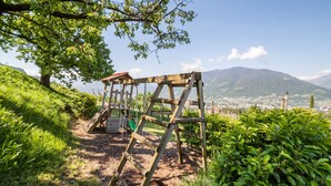 A wooden swing set in a park with a view of a town and mountains in the background. - Vacation Apartment "Sauvignon" with Mountain View, Wi-Fi, Balcony, Terrace & Garden (Meran)
