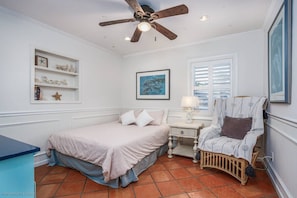 A bedroom with a bed, a chair, a nightstand, a ceiling fan, a window with blinds, and a shelf with decorative items.