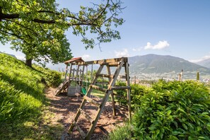 Un parque infantil con columpio de madera, con vistas a un pueblo y montañas al fondo.