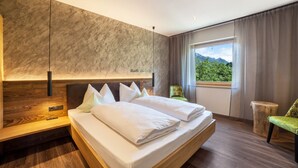 Une chambre d’hôtel moderne avec un grand lit, une tête de lit en bois, une table de chevet, un fauteuil vert et une vue sur la verdure par la fenêtre.