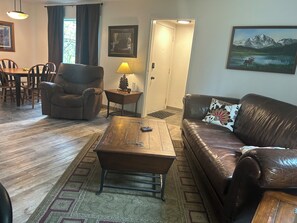 Cozy living space with a sofa, armchair, and coffee table, featuring a dining area and a door to another room.