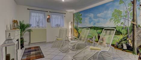 A room with a mural, three white chairs, a small table with a lamp, and a radiator.