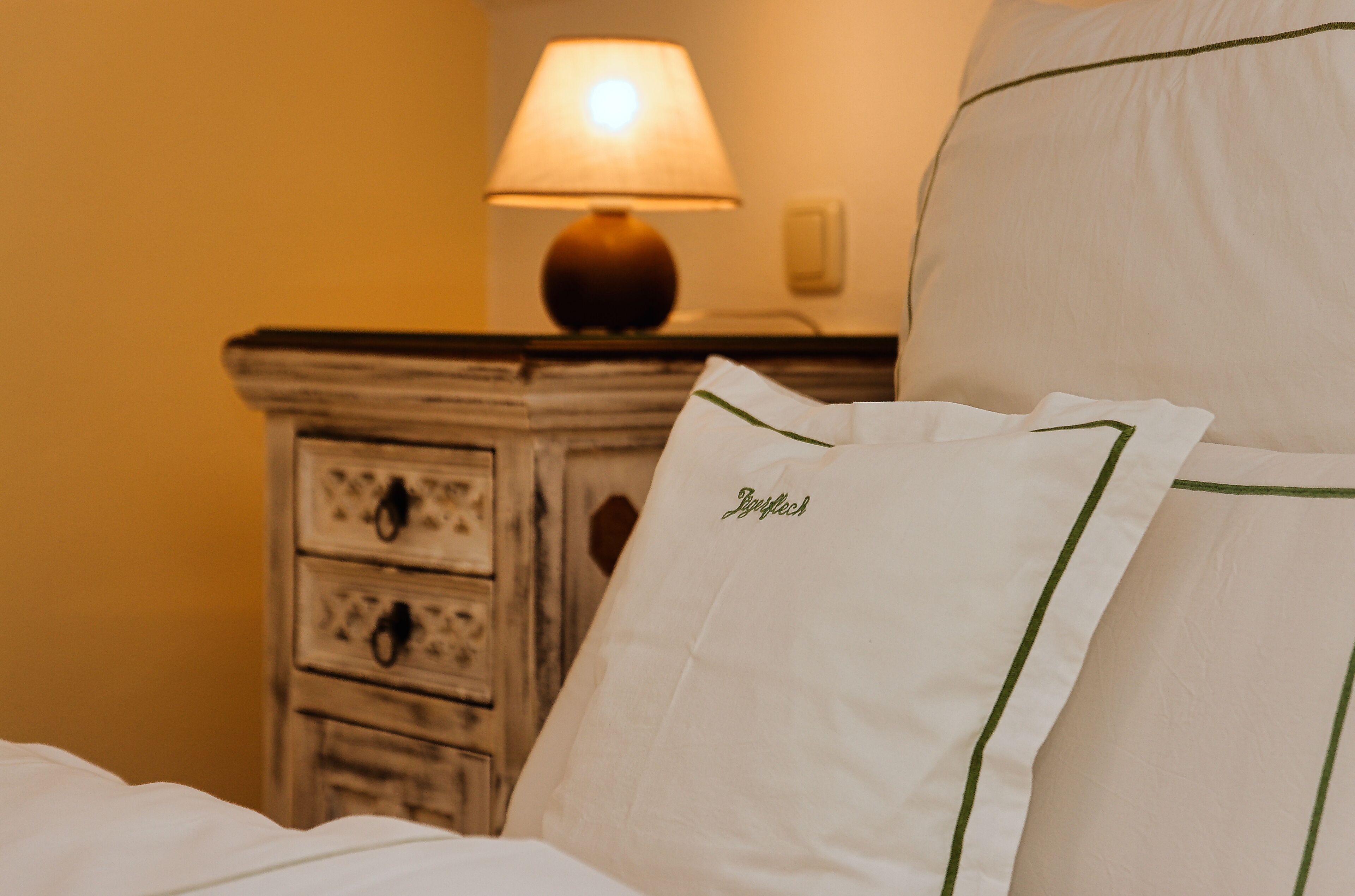 A bed with white bedding and a pillow with embroidered text, a wooden nightstand with drawers, and a lamp.
