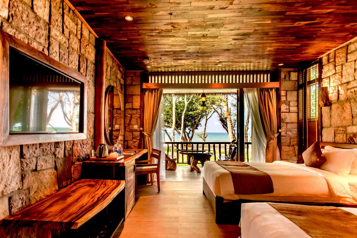 A room with a stone wall, wooden ceiling, a bed, a desk, and a view of the sea.