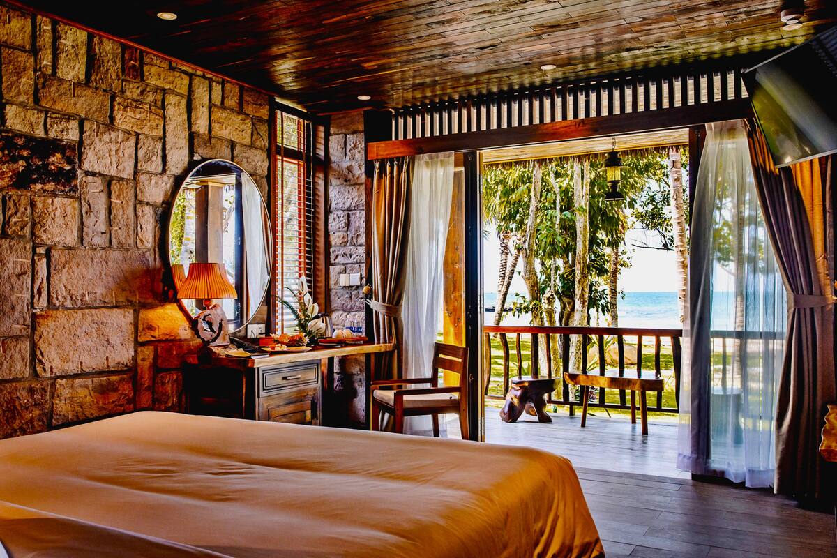 A bedroom with a large bed, a desk with a lamp, a chair, and a view of the ocean through a sliding glass door.