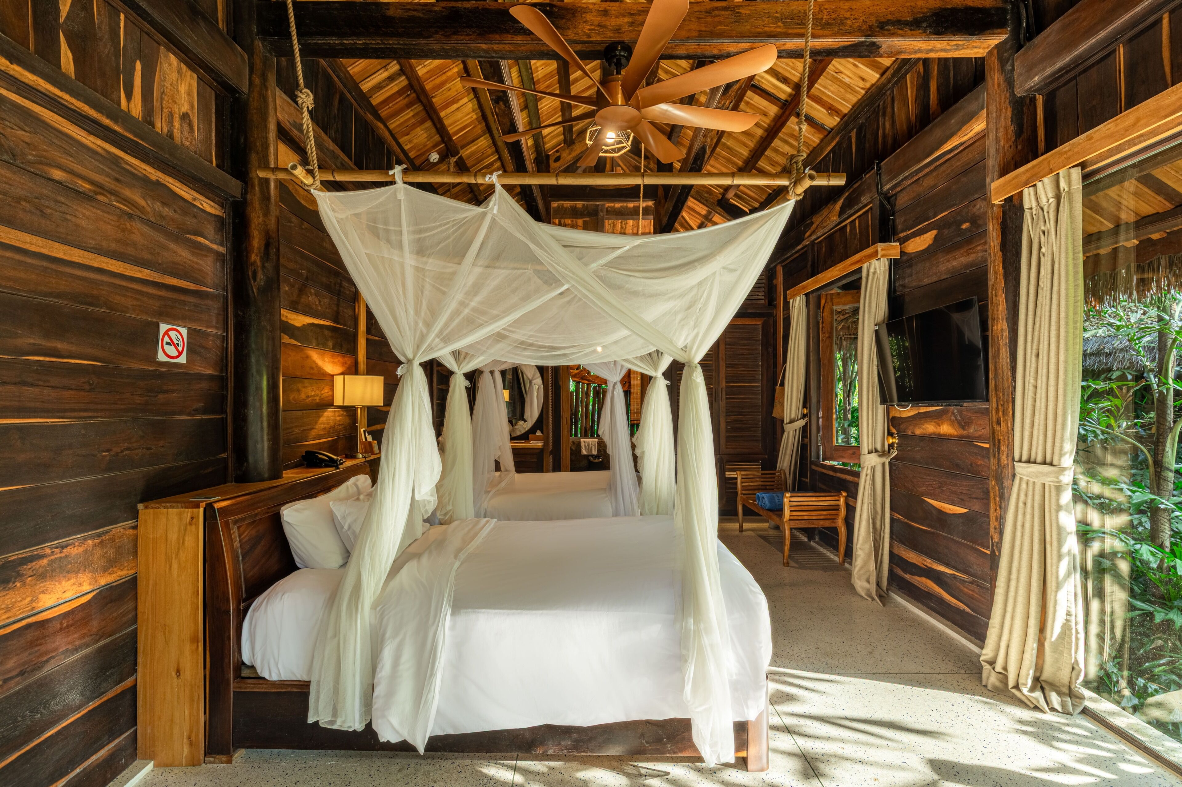 Una cabaña de madera con una cama bajo una toldo, un ventilador de techo y vistas al follaje exterior.