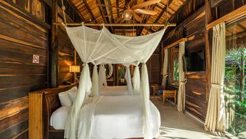 Una cabaña de madera con una cama bajo una toldo, un ventilador de techo y vistas al follaje exterior.