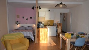 Een knus woongedeelte met een bed, een klein keukentje, een eethoek en een geel fauteuil.