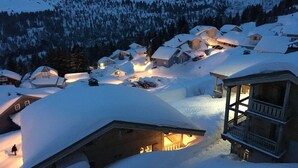 A snow-covered village with wooden houses, a wooden structure with a balcony, and a lit interior. - Cozy Mountain Stay w/ Balcony (Arâches-La-Frasse)
