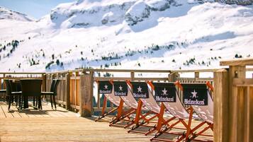 Une terrasse en bois avec des chaises longues et des tables aux couleurs de la marque Heineken, offrant une vue sur un paysage montagneux enneigé.